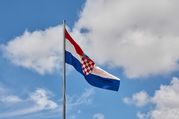 The flag of Croatia with its red and white checkerboard coat of arms waves on a flagpole. A bright blue sky with fluffy white clouds serves as background.