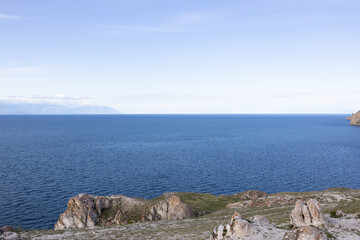 A serene view of Lake Baikal with calm waters reflecting the sky. Mountains and an island are visible in the distance under a cloudy sky.