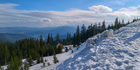 Beautiful winter scene with snow covered pine forest, cozy mountain cabin and glowing sunset over misty hills.