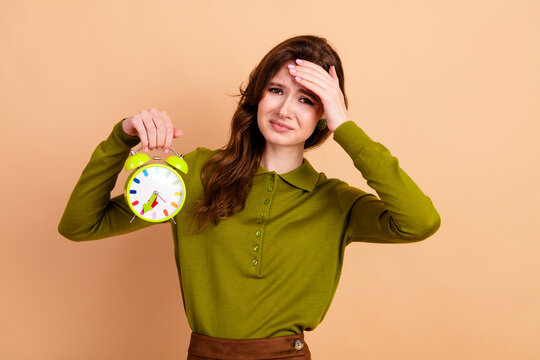 Young woman holds bright green alarm clock and looks stressed as she checks her forehead against a warm beige background conveying time pressure morning stress