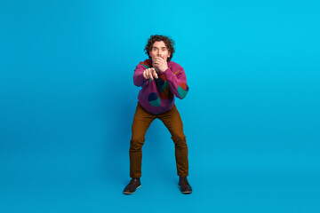 Young man in a colorful sweater points at the camera with a playful pose against bright blue background creating a casual fashion lifestyle