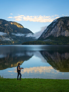 Ein Mann mit Kamera steht am Ufer eines klaren Bergsees. Vor ihm spiegeln sich Wolken und Berge im stillen Wasser einer friedlichen Morgenlandschaft.