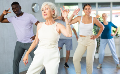 Cheerful active senior woman having fun and dancing hip hop during workout in dance class with mixed age multiracial group of adults