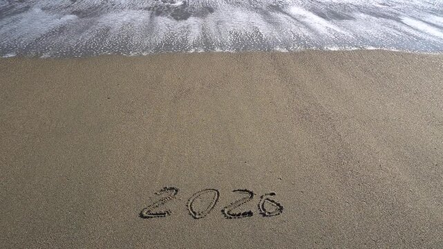 A sea wave rolls over the inscription "2026" in the sand. Surf and foam, seashore, video clip.