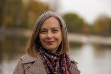 Woman with silver hair smiles serenely by the water on a cool autumn day in a park