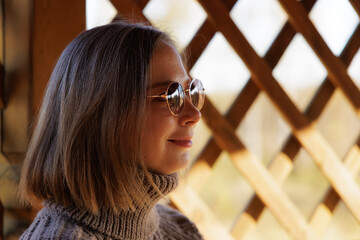 Smiling woman in round sunglasses enjoys a sunny moment near a wooden lattice backdrop