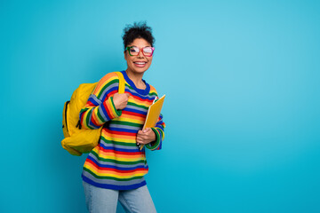 Young woman student with rainbow striped sweater and yellow backpack smiling while holding a folder against a bright blue background