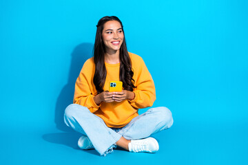 Young mixed race woman sits cross legged in a bright yellow sweater holding a phone against a vivid blue background for a modern lifestyle stock portrait