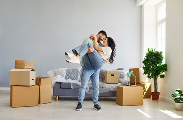 Couple in love embrace while moving home. Among boxes in living room, he lifts his partner, celebrating moving day and unpacking as they start family life. Real estate, move and rent concept. 