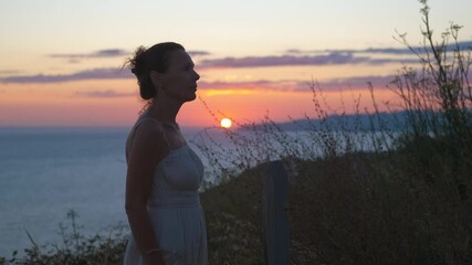 Silhouette of a pensive woman admiring a beautiful seaside sunset. Solitary woman standing in profile on a cliff, enjoying the beautiful sunset over the calm ocean, representing a moment of peace