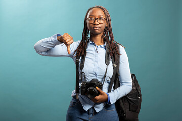 Unimpressed photography enthusiast makes a thumbs down sign while posing with dslr camera against isolated background. African american female photographer showing a disapproval gesture.