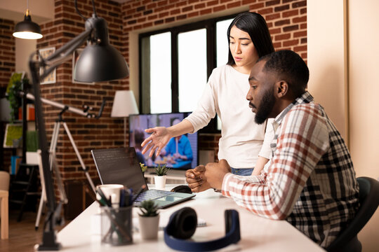 Supportive girlfriend discussing software on laptop with focused male developer at home office desk. Multiethnic couple working remotely together and brainstorming on app development for tech company.