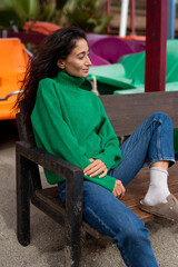 Woman in a green turtleneck sweater and jeans sitting on a wooden bench by the beach, enjoying the breeze on a peaceful autumn day.