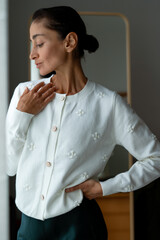 Elegant woman in a white embroidered sweater with pearl buttons standing indoors, touching her neck gently and looking down with calm expression in soft daylight