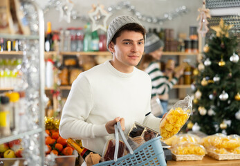 Male shopper choosing food and drinks in supermarket for christmas celebration. In background, female salesperson decorates a Christmas tree with toys