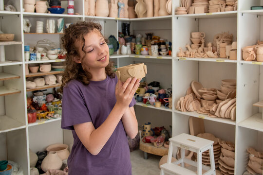 Close-up portrait of a smiling young girl in an apron in a pottery studio. Joyful child surrounded by unfired clay ceramics, emphasizing creative learning and hands-on craft. - Powered by Adobe