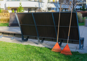 Two orange leaf rakes leaning against a modern outdoor bench in a landscaped residential courtyard.