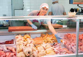 In meat department, saleswoman and disposable gloves offers to buy turkey fresh poultry. Butcher shop female clerk points with hand at fresh large juicy pieces of chicken in refrigerator window