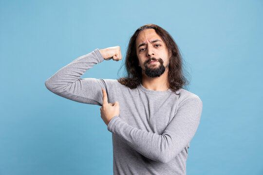 Strong fit guy pointing at his flexed muscles in front of the camera, showing off his toned athletic physique against blue background. Physical wellness and determination serve as fitness motivation.