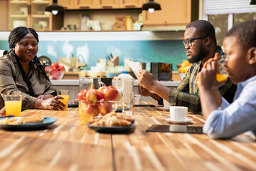 African american dad reading daily newspaper at the kitchen table, enjoying morning routine during breakfast with his little family. Young dad feeling happy chatting with his children and wife, eat