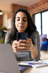 Fototapeta premium Professional woman working from home switches from laptop to mobile phone to catch up on messages and notifications. Indian female entrepreneur balances digital tasks during remote workday.