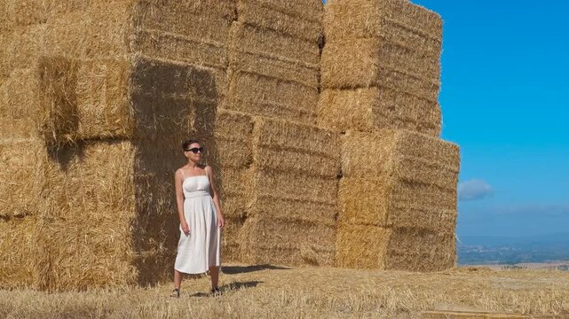 Elegant woman in white dress posing by haystacks. Stylish woman in a white summer dress and sunglasses standing confidently next to a large stack of golden hay bales in a sunny rural field