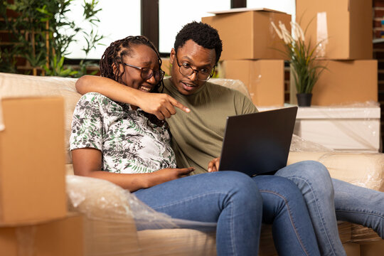 African American boyfriend and girlfriend relaxing on couch, laughing and shopping online for home furniture. Happy black couple using laptop and enjoying bonding time in new apartment.