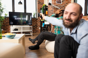African American male gamer hand on head, reacts with frustration after losing video game. Caucasian man holding wireless controller gestures dramatically while speaking with friend near television. © DC Studio