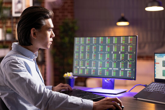 Dedicated remote worker monitoring company investments and financial graphs across multiple devices. Asian man focused on analyzing stock trading session during evening hours at home.