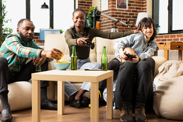 Friends enjoy gaming competition in cozy brick wall apartment. Black and white women play with wireless controllers as african american man eats chips and caucasian man sits nearby with girlfriend.