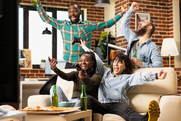 Group of friends drink and celebrate during cozy home hangout. Multicultural gaming buddies with gamepads and beer bottles cheer together, looking at television after video game win in living room.