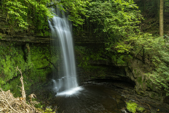 The Glencar Waterfall plunges over the moss-covered, layered cliff face into a dark pool below, surrounded by dense, lush green forest.