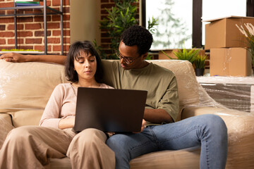 Couple sits on couch with laptop in living room surrounded by moving boxes, browsing online...