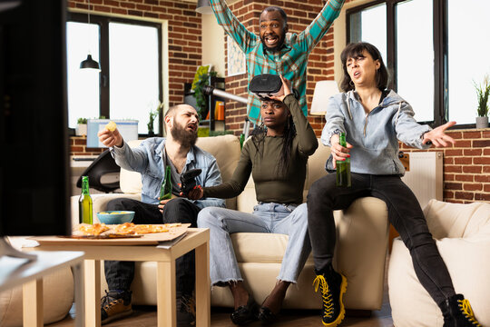 Black woman lifts AR headset, reacting angrily at TV screen after losing virtual reality game. Diverse male and female peers in living room express frustration and disappointment over 3D gaming loss.