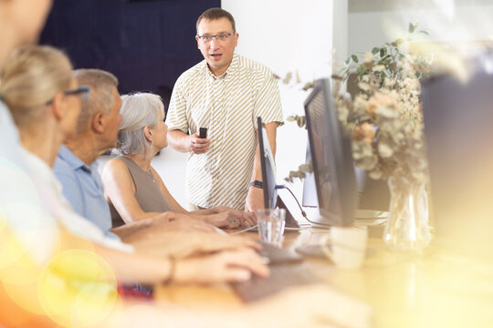 Positive friendly adult man, professional teacher, conducting group lesson of basics of computer skills to interested older men and women sitting at table