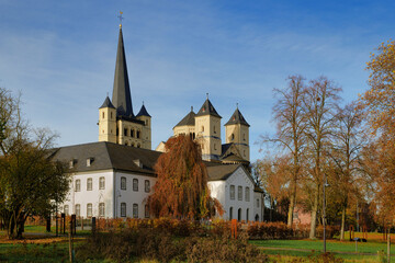 The historic Brauweiler Abbey near Cologne, featuring its Romanesque church with spires and baroque white wing, surrounded by colorful autumn foliage under a clear sky