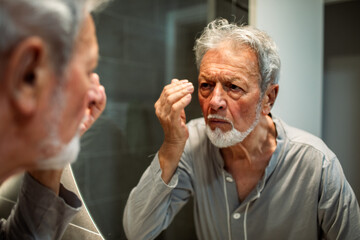 Senior man examining wrinkles with concern in bathroom mirror