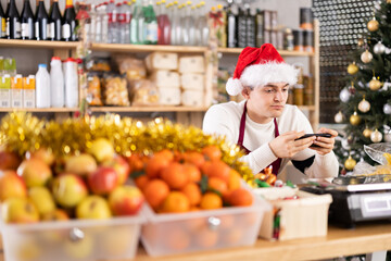 Young guy seller with phone bored sitting at counter in grocery store decorated for christmas