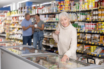 Mature woman buyer carefully chooses frozen food in grocery store freezer