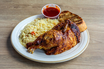 A half roast chicken with crispy skin , leg and breast, with rice and dipping sauce and a slice of garlic bread toast on a wooden table diner style