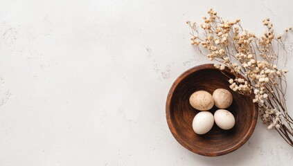 A rustic wooden bowl holds three speckled eggs and one plain white egg, nestled beside dried beige floral sprigs on a light textured background.  Plenty of copy space is visible to the left