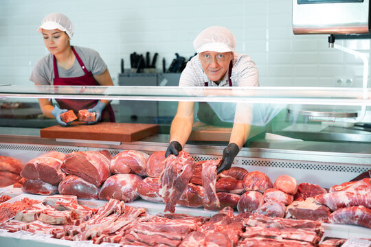 Adult man and young woman sellers in uniform display raw meat beef in butcher shop..