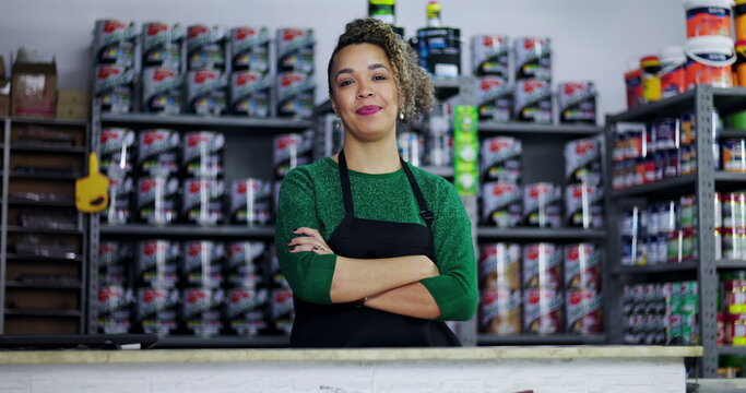 Confident young woman with curly hair standing behind a counter in a hardware store, arms crossed, smiling proudly, paint cans displayed on shelves in the background