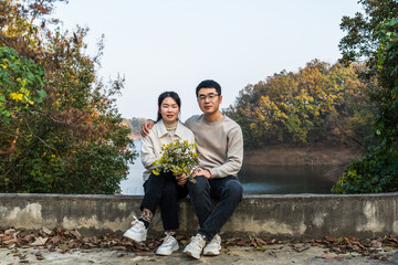 A couple sits by the lake with autumn foliage surrounding them, holding flowers in a peaceful outdoor setting.