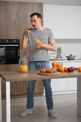 Young man with glass of orange juice eating doughnut in kitchen