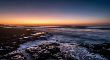 A serene coastal scene at sunset with waves crashing against rocky shoreline and a vibrant sky transitioning from orange to deep blue
