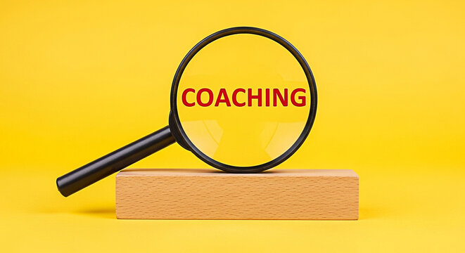Magnifying glass focusing on the word coaching displayed in red text against a bright yellow background with a wooden block stand