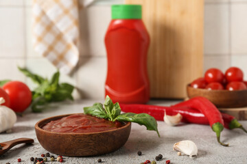 Wooden bowl of tasty ketchup with basil leaves, garlic and peppercorns on grey table
