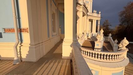 Grand curved staircase of a pale blue and white palace exterior bathed in warm golden light contrasting with a dramatic, dark storm sky at sunset or sunrise