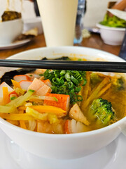 Vertical close-up of delicious Asian noodle soup. Includes vibrant crab sticks, fresh broccoli, carrots, green onions. Chopsticks ready for a hearty meal